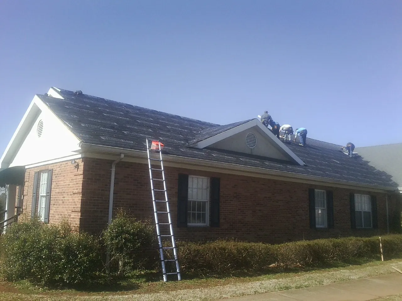 Crew tearing shingles off a brick ranch roof with a ladder set — Emergency Roof Repair work by our crew in Howe