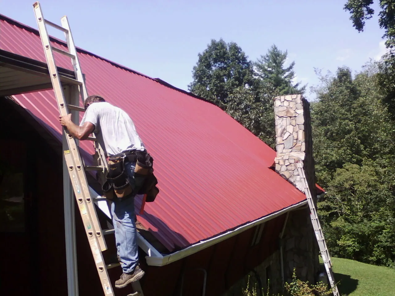 Roofer on a ladder beside a red metal roof and stone chimney — Soffit & Fascia Repair by Roofing Brothers in Howe